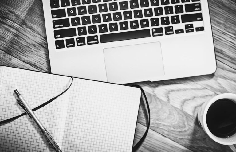 black and white image showing a bird's eye view of a laptop keyboard and an open notebook with a pen